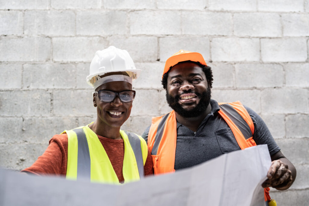 A black man and a black woman wearing hard hats and hi-vis, looking at construction plans