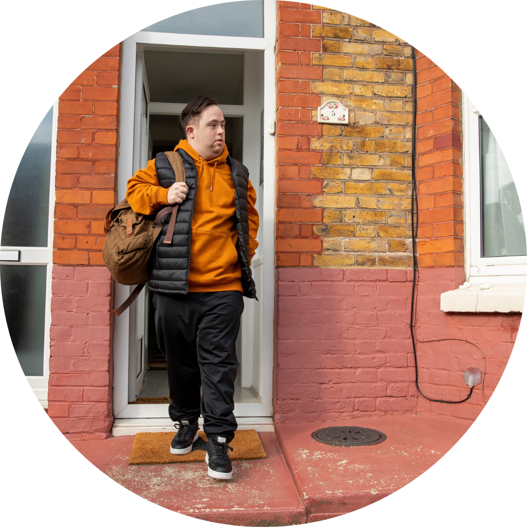 A young man who has down syndrome leaves a red brick housing with a black backpack, organge hoodie and trainers