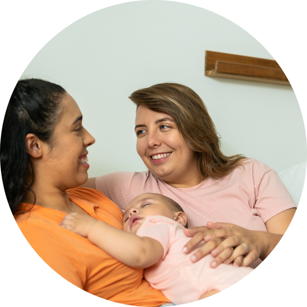 A couple - two women, one white and one black sit together on a sofa, enjoying time together while baby sleeps in their arms