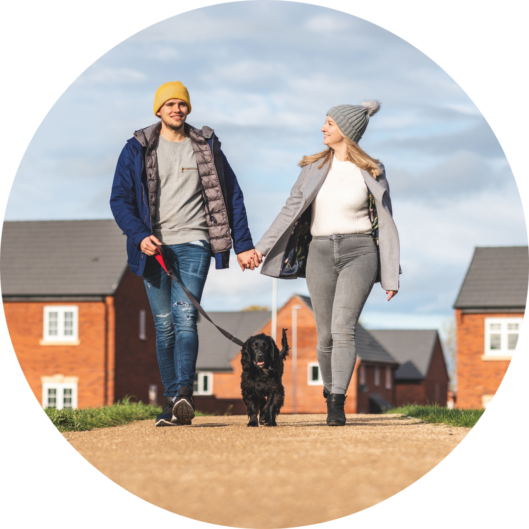 A young smiling couple (man and woman) walk a black dog on a leash on a sunny autumn day, with blurred views of a red brick housing estate in the background