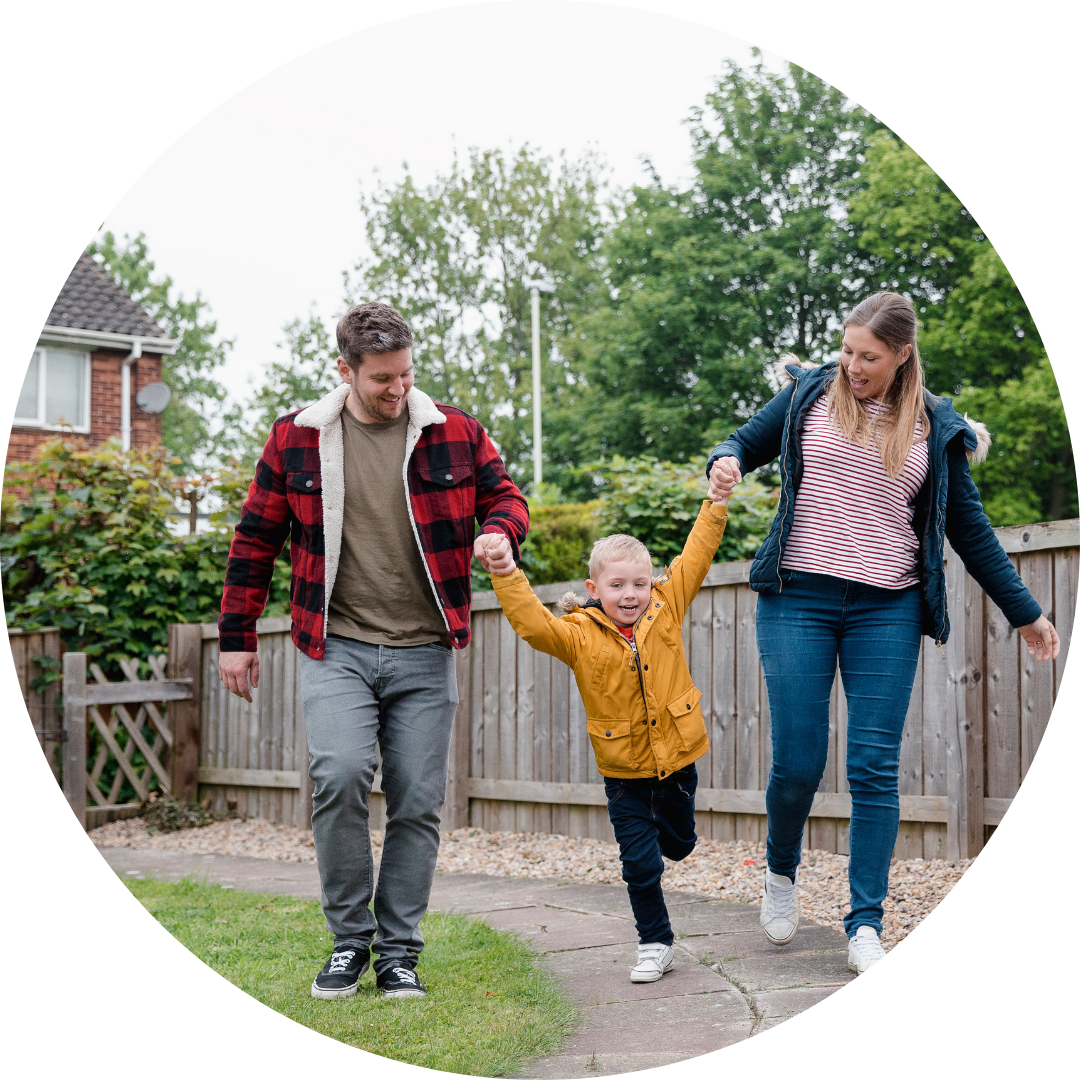 Little boy is in between his parents (a man and a woman) holding their hands as they walk along the garden path to get back to the house. They are smiling and laughing on the way back.