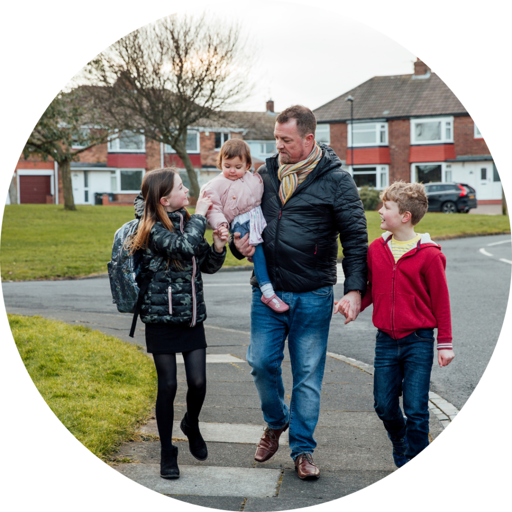 Mature man has just picked up his eldest daughter from school with his other two children. They are talking as they walk home together. The man is carrying the youngest child in his arms. They walk in front of a rown of semi-detached houses