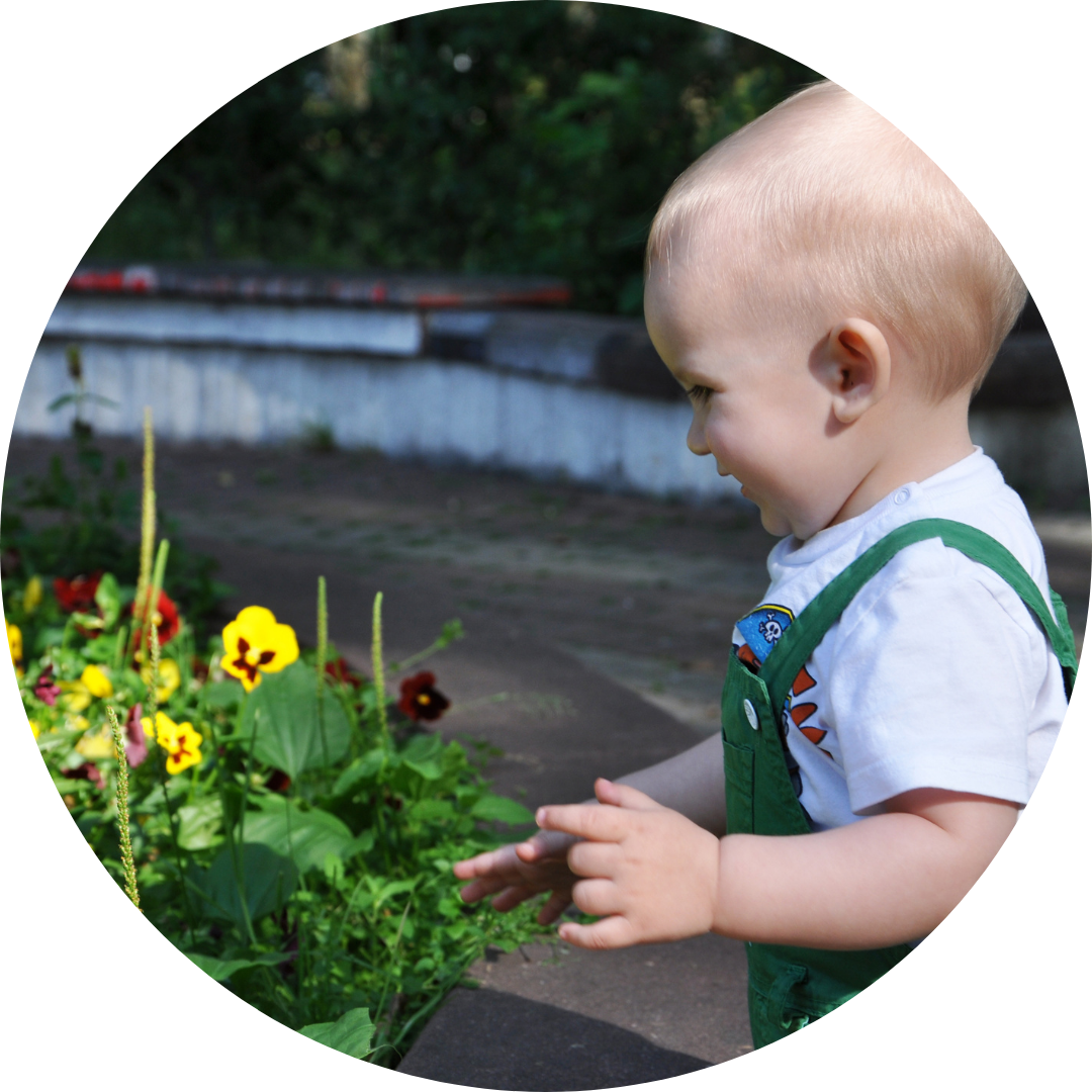 A young toddler with blonde hair and green dungarees stands smiling in front of a pot of yellow and red flowers in a garden