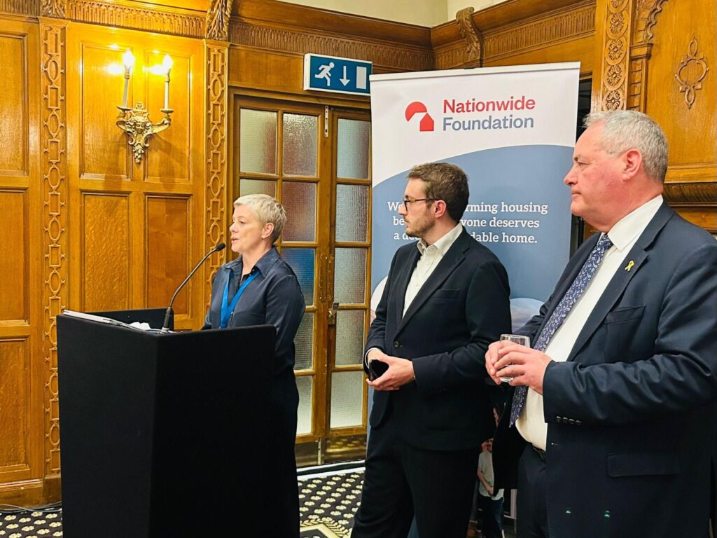 A woman stands behind a lecturn speaking to a room we can't see. Next to her, two men stand in suits