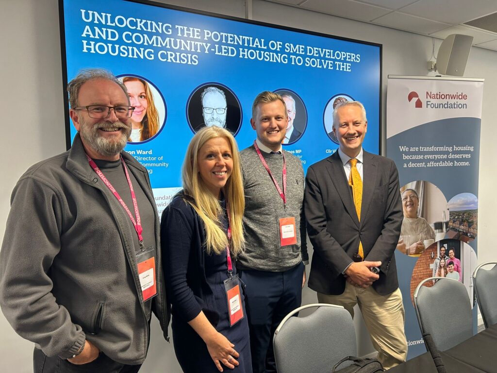 Four people - three men and a woman stand in a line smiling at the camera. Behind them a secreen shows an image of their faces with the title 'Unlocking the potential of SME developers and community-led housing to solve the housing crisis'