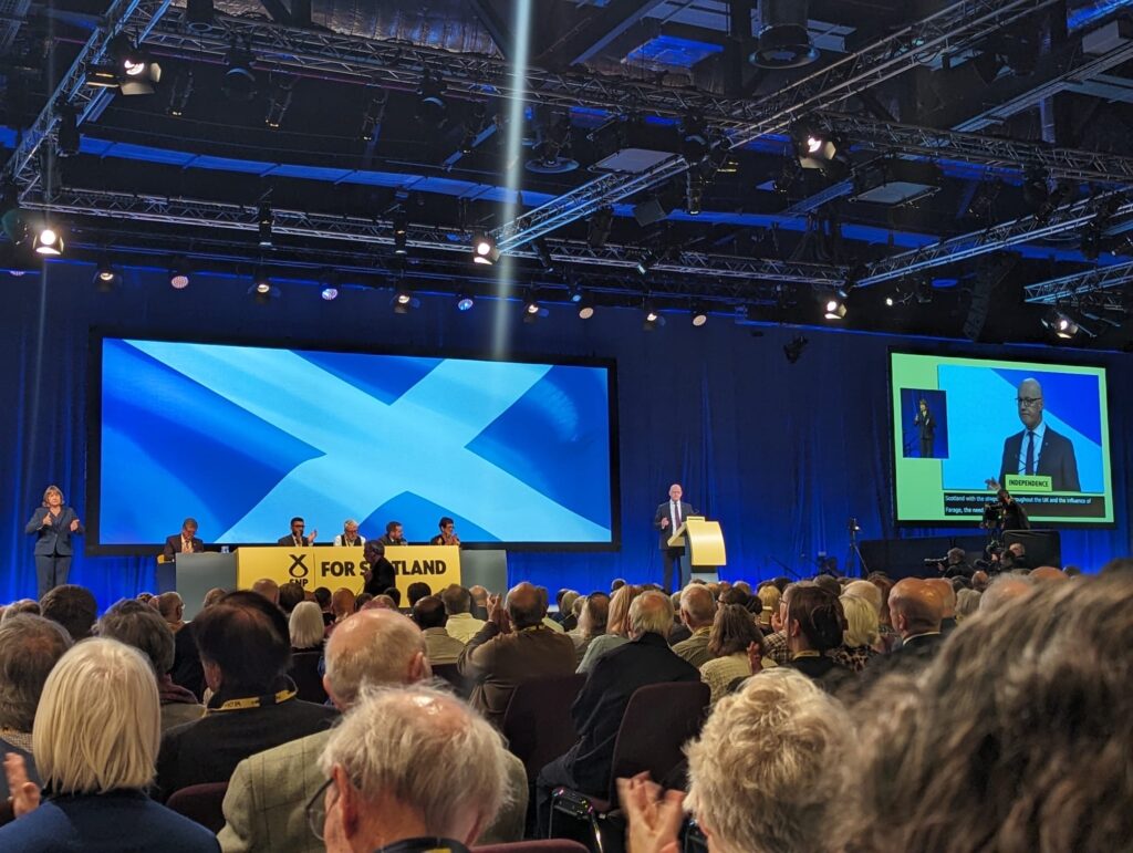Image taken from the busy crowd at a conference looking at a stage of people sat behind a table and one man at a lecturn. Behind them is a very large image of the scottish flag